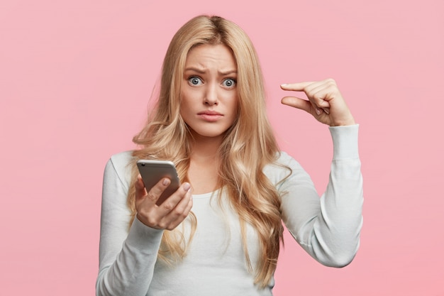 Displeased female model holds cell phone, shows something very little with hands, uses free internet connection, isolated over pink wall. People, facial expressions and technology concept