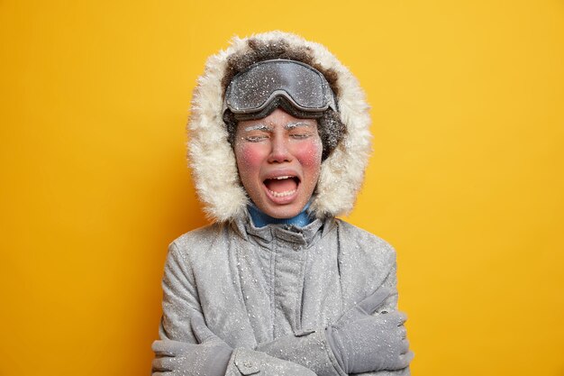 Displeased frozen girl in winter clothing trembles from cold and embraces herself expresses negative emotions has red face covered with hoarfrost.