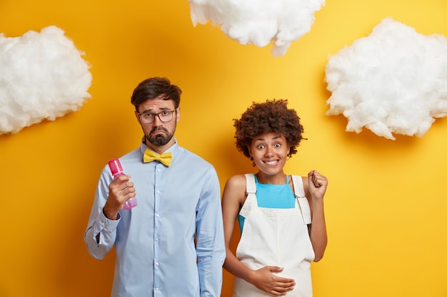 Displeased future father holds feeding bottle, supports expectant wife, prepare for child birth, isolated on yellow . mom expects baby holds pregnant tummy caressing belly. maternity concept