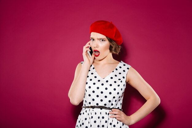 Displeased ginger woman in dress with arm on hip talking by smartphone and looking at the camera over pink