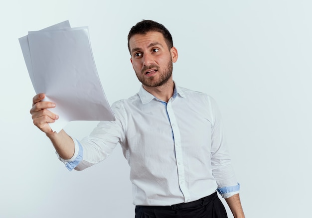 Free photo displeased handsome man holds and looks at paper sheets isolated on white wall