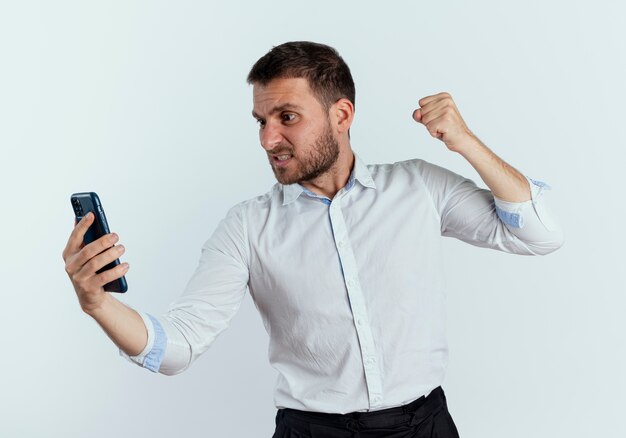 Displeased handsome man keeps fist ready to punch looking at phone isolated on white wall