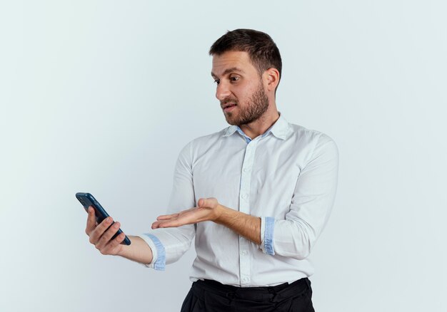 Displeased handsome man looks and points at phone isolated on white wall