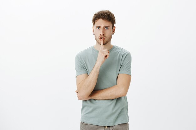 Displeased handsome serious employer in casual t-shirt, frowning and saying shh, demanding silence