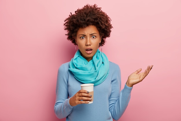 Foto gratuita donna dalla pelle scura indignata scontenta con l'acconciatura riccia, alza la mano, guarda con frustrazione, vestita con abiti blu casual, tiene il caffè da asporto isolato sopra il muro rosa. emozioni negative