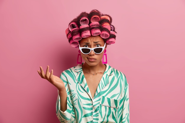 Displeased indignant woman wears curlers for getting hair styled, raises hand and puzzled by unpleasant news, wears stylish sunglasses and dressing gown, stands indoor against pink wall