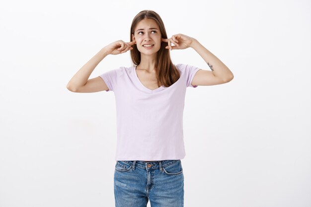 Displeased intense bothered female with tattoo clenching teeth closing ears with index finger looking at upper right corner interrupted with loud sound