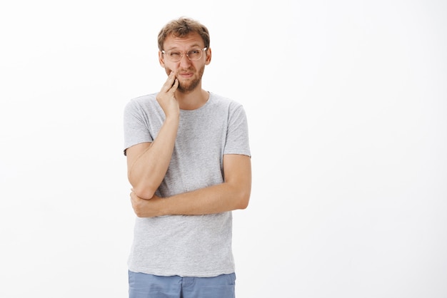 Displeased intense charming european male with bristle squinting and wrinkling nose touching cheek while feeling painful toothache having decay or dental problems