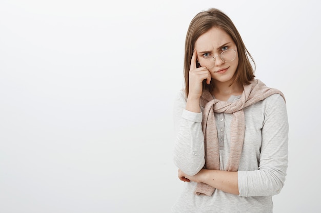 Displeased intense and tired female tutor in glasses frowning looking from under eyeglasses holding finger on temple thinking person is dumb losing temper from irritation and annoyance over grey wall