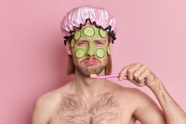 Displeased man applies green nourishing mask on face with cucumber slices to rejuvenate skin looks sadly at camera brushes teeth stands topless indoor against pink background.