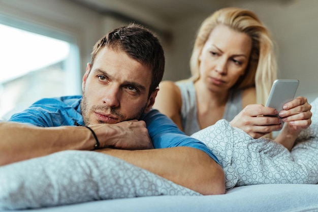 Бесплатное фото Недовольный мужчина лежит на кровати и игнорирует свою девушку, которая отправляет текстовые сообщения на мобильный телефон