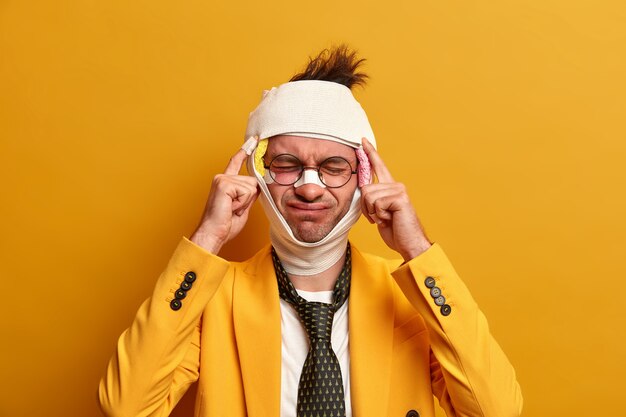 Displeased man suffers from unbearable migraine after injury, dressed in formal clothes, has bruises and broken nose, recovers after difficult surgery operation, isolated on yellow wall