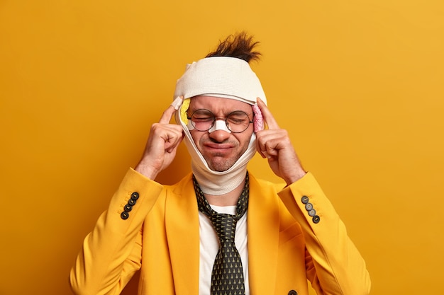 Бесплатное фото Недовольный мужчина страдает от невыносимой мигрени после травмы, одет в формальную одежду, имеет синяки и сломанный нос, выздоравливает после сложной хирургической операции, изолирован на желтой стене