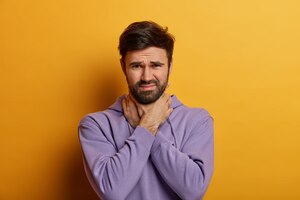 Displeased man suffocates because of painful strangle in throat, touches neck, looks dissatisfied , has sore throat after catching cold, dressed casually, poses over yellow wall