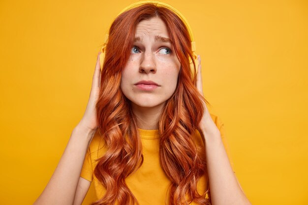 Displeased melancholic ginger teenage girl keeps hands on stereo headphones thinks about somethig sad while listening music dressed casually looks aside sadly 