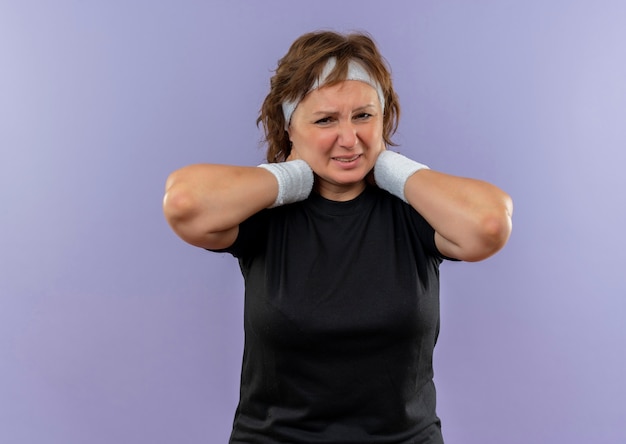 Displeased middle aged sporty woman in black t-shirt with headband looking unwell touching her neck feeling pain standing over blue wall