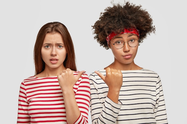 Foto gratuita le amiche dispiaciute di razza mista litigano, si puntano l'un l'altra, stanno spalla a spalla, provano malcontento dopo il litigio, vestite con maglioni a righe, isolate su un muro bianco