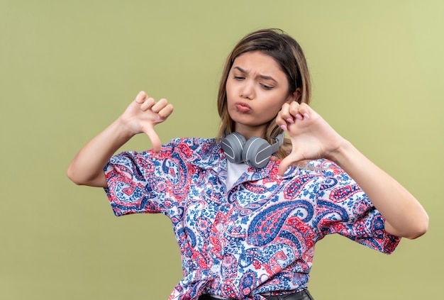 A displeased pretty young woman in paisley printed shirt wearing headphones showing thumbs down on a green wall