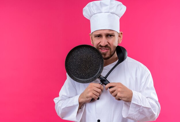 不機嫌なプロの男性シェフが白い制服で調理し、ピンクの背景の上にスープ鍋とパン立って保持している帽子を調理します。