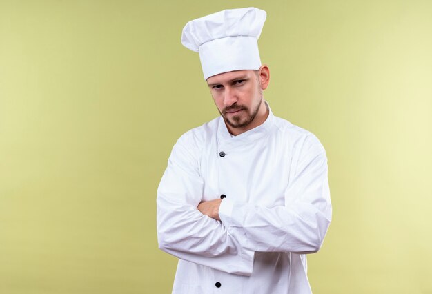 불쾌한 전문 남성 요리사가 흰색 유니폼을 입고 요리사 모자 서 팔을 넘어 녹색 배경 위에 자신감을 찾고 넘어