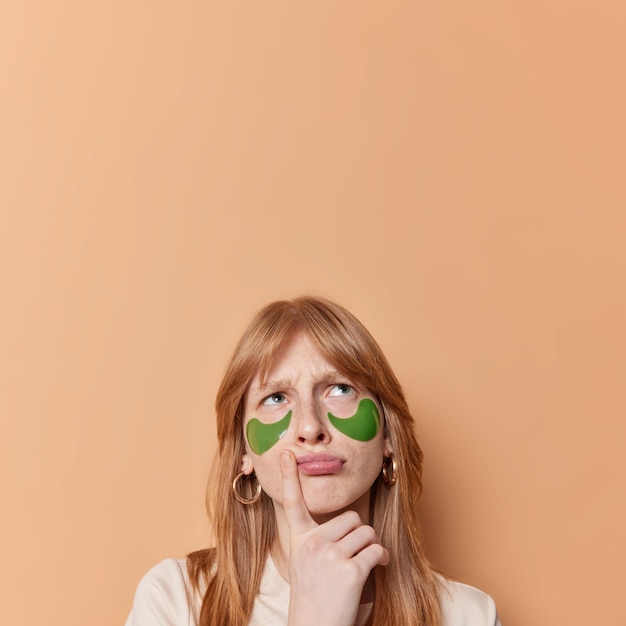 Displeased red haired girl frowns and looks above keeps finger near lips thinks about something applies beauty patches for skin care isolated over brown background blank copy space overhead