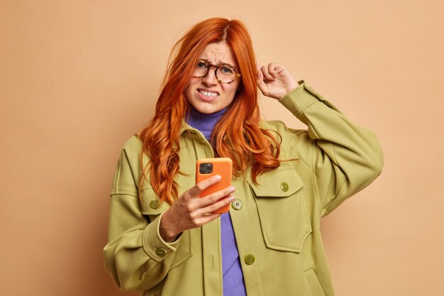 Недовольная рыжеволосая женщина чешет голову, хмурится от неудовлетворенности, пытается решить проблему со смартфоном, не знает, как использовать новое приложение, одетая в модную одежду.