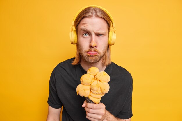 Displeased redhead man frowns face makes unhappy grimace raises eyebrows has dirty face while eating ice cream wears wireless stereo headphones for listening music isolated over yellow  wall