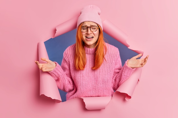 Displeased redhead woman going to cry spreads palms and has doubtful expression feels miserable wears hat and knitted jumper.