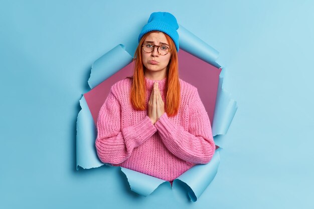 Displeased redhead woman presses palms together looks with imploring pleading face expression asks for favor purses lips had sad look wears blue hat and knitted sweater.