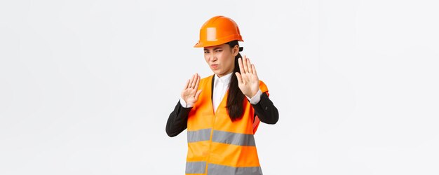 Displeased and reluctant asian female architect in safety helmet grimacing from aversion showing stop gesture refuse or rejecting awful disgusting plan standing disappointed white background