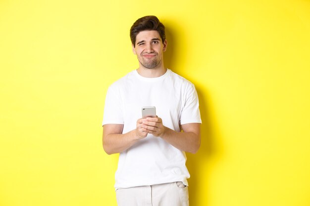 Displeased and reluctant man grimacing, being unamused by message on smartphone, standing over yellow background