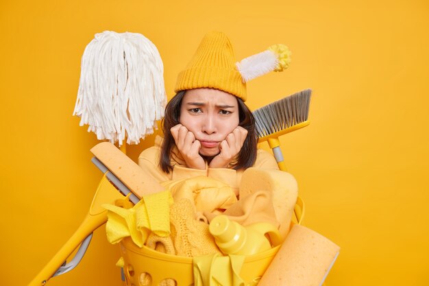 不機嫌な悲しい女性は部屋を掃除したくない混乱を悲しげに見て、汚れは黄色い壁に対して洗濯かごの近くでさまざまな掃除道具のポーズを使用します