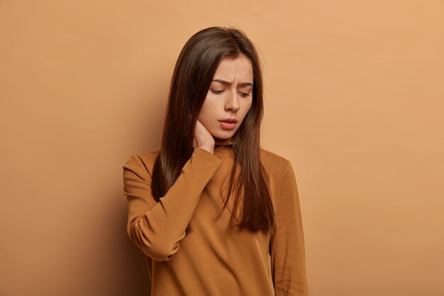 Displeased sad woman touches neck, looks down with unhappy expression, thinks about her problems with worried look, wears brown jumper