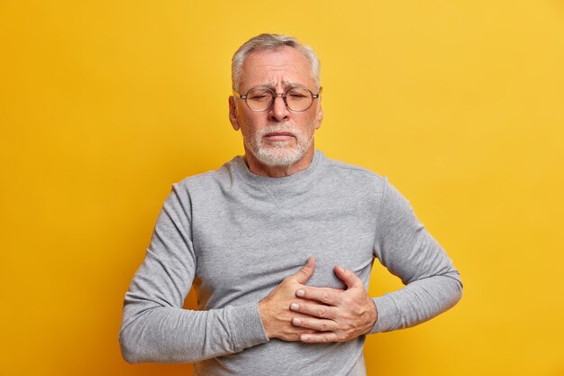 Displeased senior man presses hand to chest has heart attack needs pinkillers dressed in casual turtleneck wears spectacles isolated over yellow wall