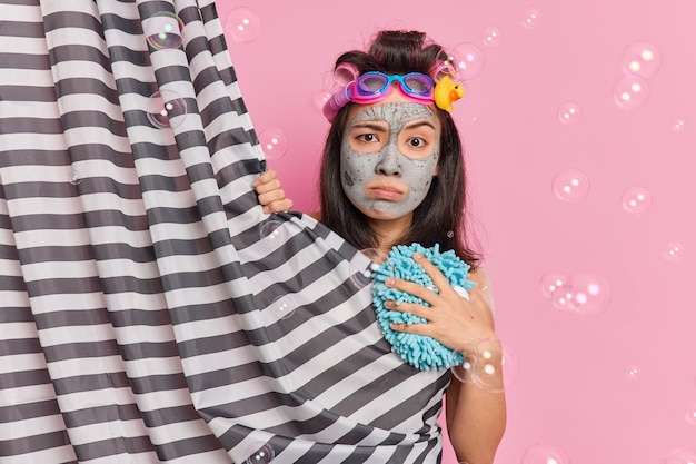 Free photo displeased serious asian woman looks directly at camera hides behind shower curtain undergoes beauty procedures in douche applies clay facial mask poses against pink background with bubbles around