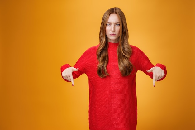 Displeased serious-looking female in knitted red dress frowning looking concerned as pointing down at displeasing unappropriate thing standing disappointed and unhappy over orange background