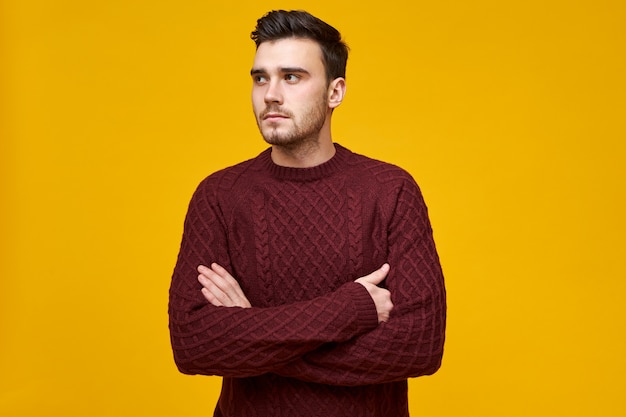 Displeased serious young brunette guy in stylish knitted pullover posing , crossing arms on his chest