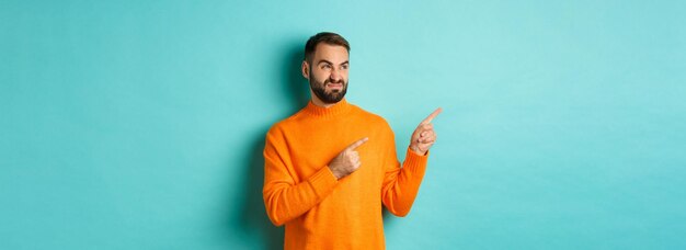 Foto gratuita ragazzo dispiaciuto e scettico che fa una smorfia mostrando un cattivo prodotto che punta le dita a sinistra con delusione