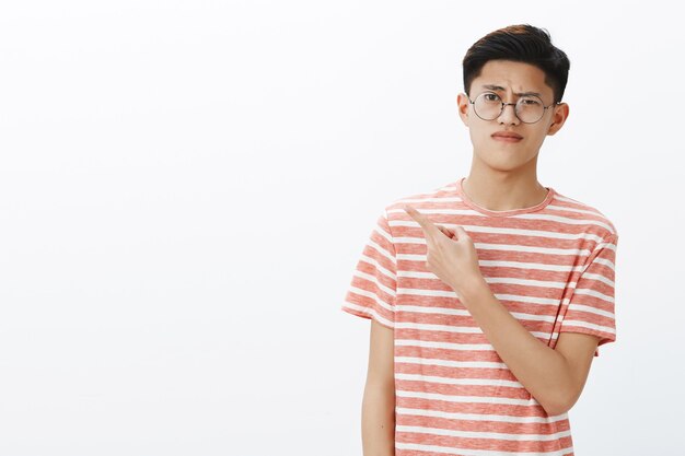 Displeased smart young attractive asian male student in glasses and t-shirt raising eyebrow in scorn and doubt pursing lips from dislike pointing backwards or left questioned