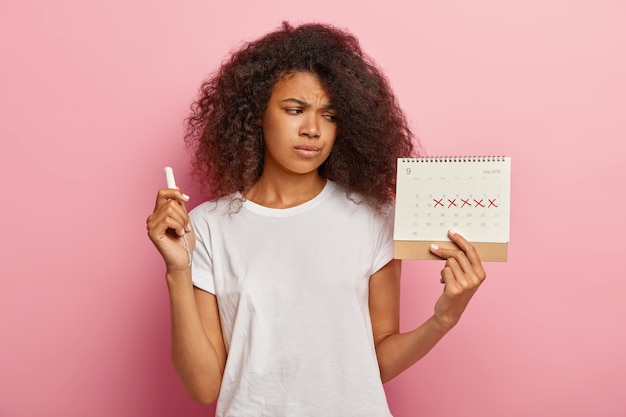 Недовольная стрессовая темнокожая женщина смотрит на календарь месячных с отмеченными красными крестами