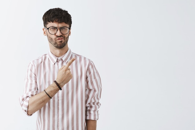 Displeased stylish bearded guy posing against the white wall with glasses