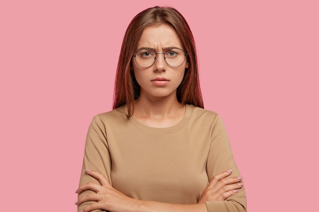 Displeased sullen female has discontent expression, keeps hands crossed, shows her dislike, frowns face, feels unhappy, has staight dark hair, poses against pink wall. Negative emotions concept