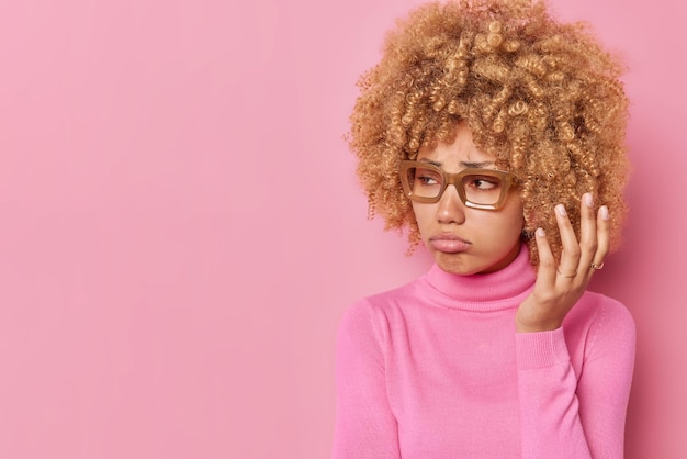 Displeased sullen young woman looks away with upset expression wears transparent glasses casual turtleneck expresses negative emotions poses against pink background copy space for your text