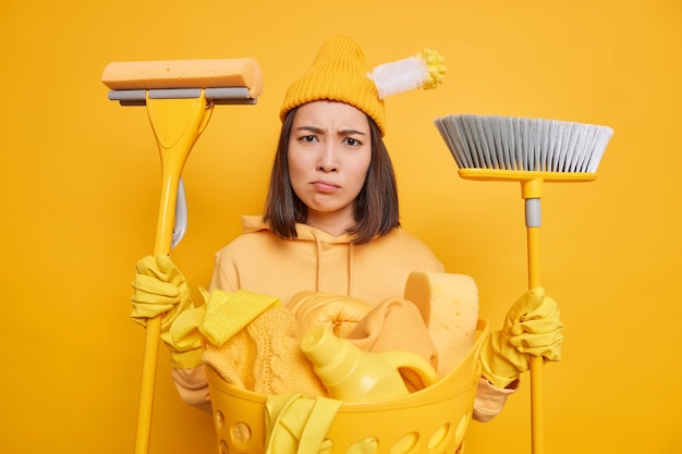 무료 사진 불쾌한 피곤한 아시아 여성이 얼굴을 찡그린 얼굴은 집을 청소하려는 욕망이 없습니다. 걸레와 빗자루는 세탁을 하느라 바쁘게 착용하고 보호 고무 장갑 모자와 노란색 배경 위에 절연 운동복을 착용합니다.