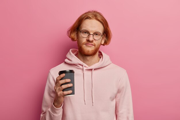 Displeased tired student tries to refresh with strong cup of takeaway coffee, looks with upset expression , dressed in hoodie, needs good rest, has ginger bob hair isolated on pink wall