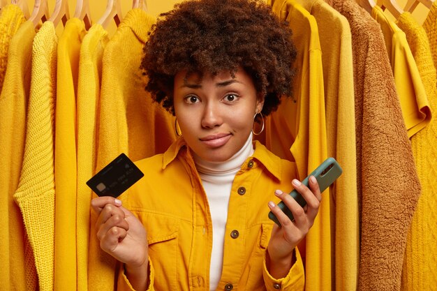 Displeased unaware woman with curly hairstyle, unable to pay all sum of money for clothes, holds plastic card and modern mobile phone, poses against plain yellow jumpers on hangers.