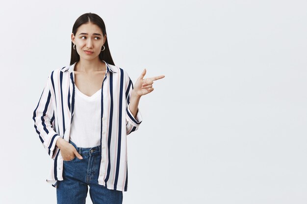 displeased unimpressed stylish female in striped blouse and jeans, frowning and smirking while looking and pointing right, being doubtful while posing over gray wall