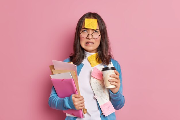 Displeased university student bites lips and looks above with unhappy tired expression drinks takeaway coffee works with papers wears round spectacles.