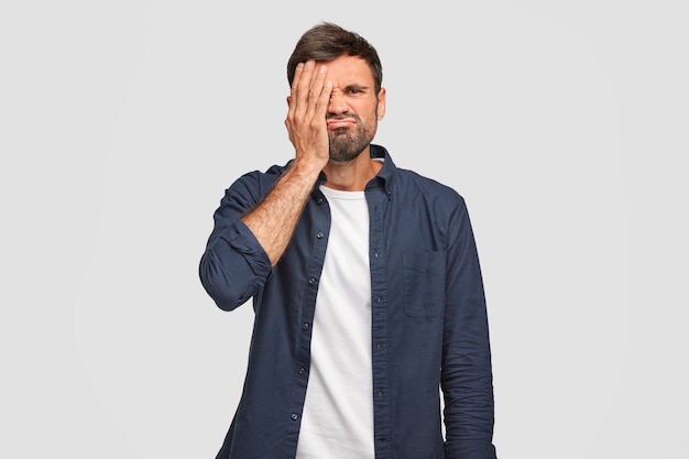 Displeased unshaven male has bothered face, covers eye with hand, feels bored, frowns, dressed in dark blue fashionable shirt, stands against white wall. People and facial expressions.