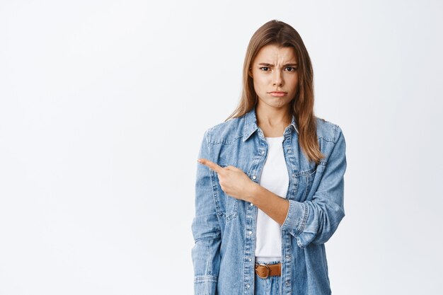 Displeased and upset young woman frowning offended and complaining, pointing finger left at white empty space, standing against studio wall